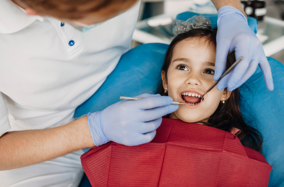A dentist examines a young girls teeth as she sits in a dental chair with a red bib.