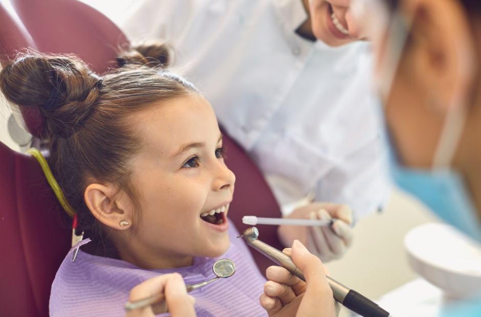 Young girl smiling in a dentist’s chair while dentists prepare dental tools for her checkup.