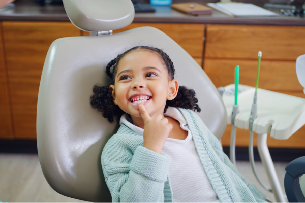 Smiling young girl sits in a dentist chair, pointing to her teeth in a dental clinic.
