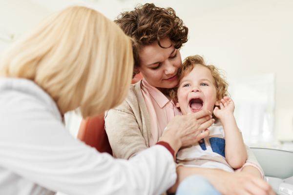 A child sits on a woman’s lap while a doctor checks the child’s mouth in a clinic.