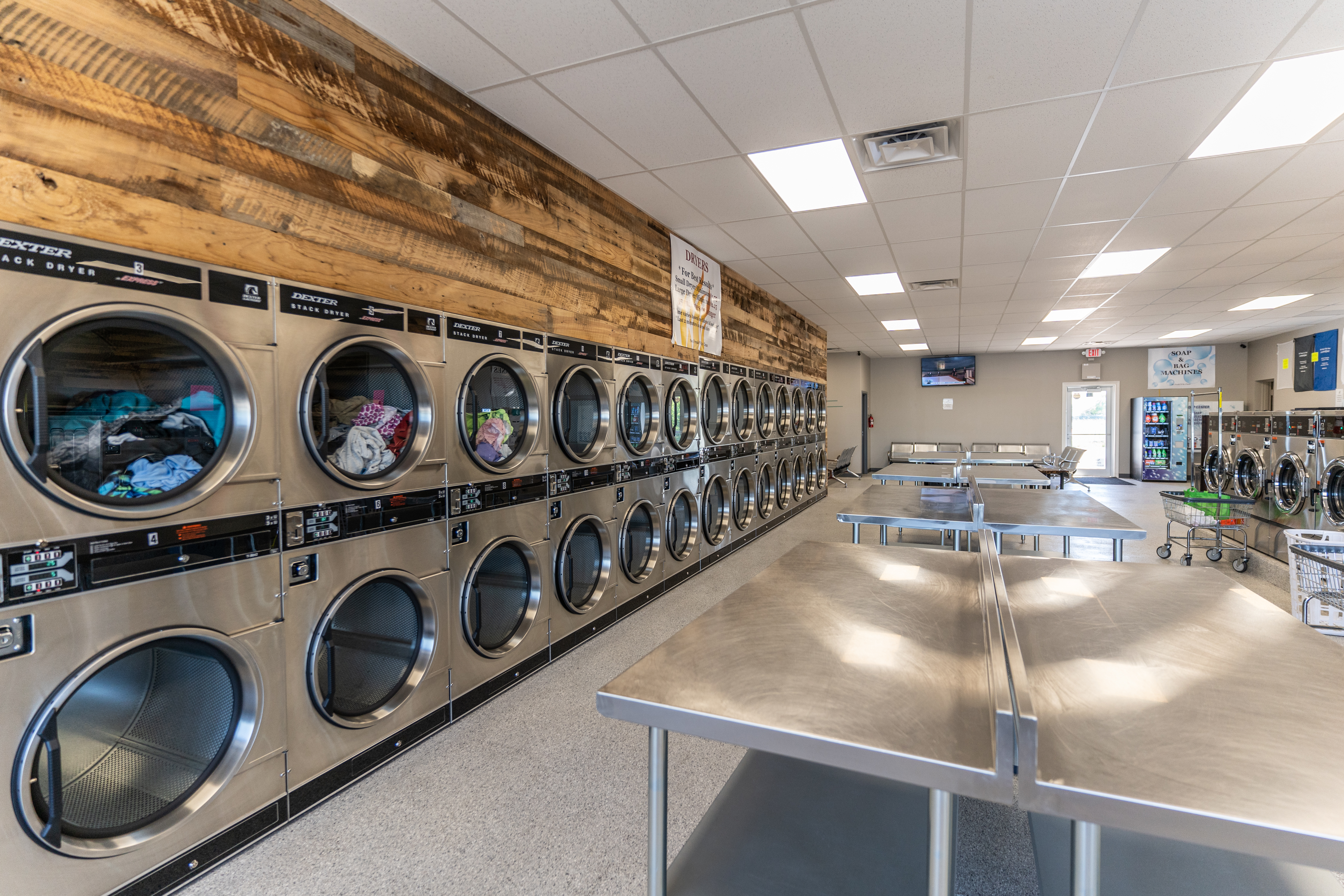 Interior of a laundromat with rows of industrial washers, folding tables, and bright lighting.