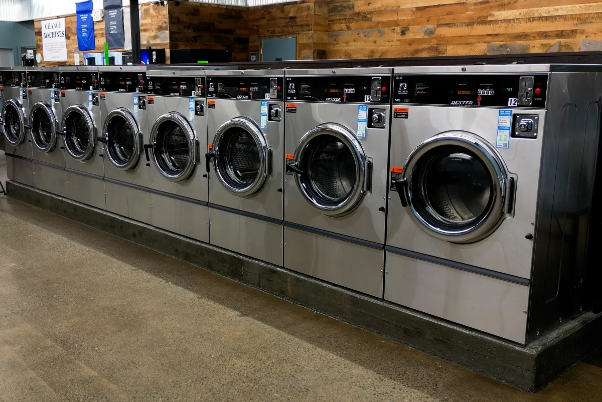 A row of stainless steel washing machines in a laundromat with a polished concrete floor.