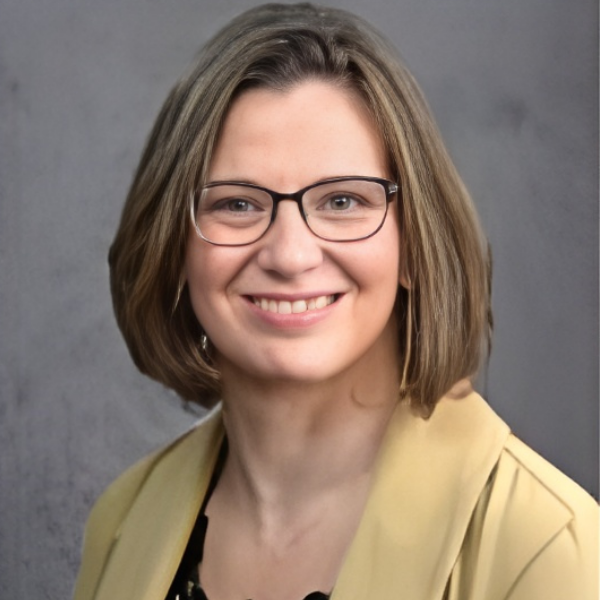 Smiling woman with glasses and short brown hair wearing a beige blazer against a gray background.