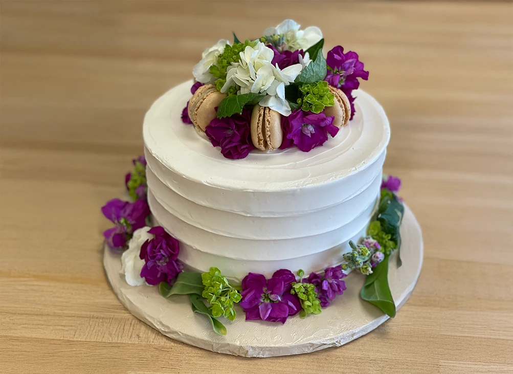 A white frosted cake decorated with macarons and colorful fresh flowers on top and around.