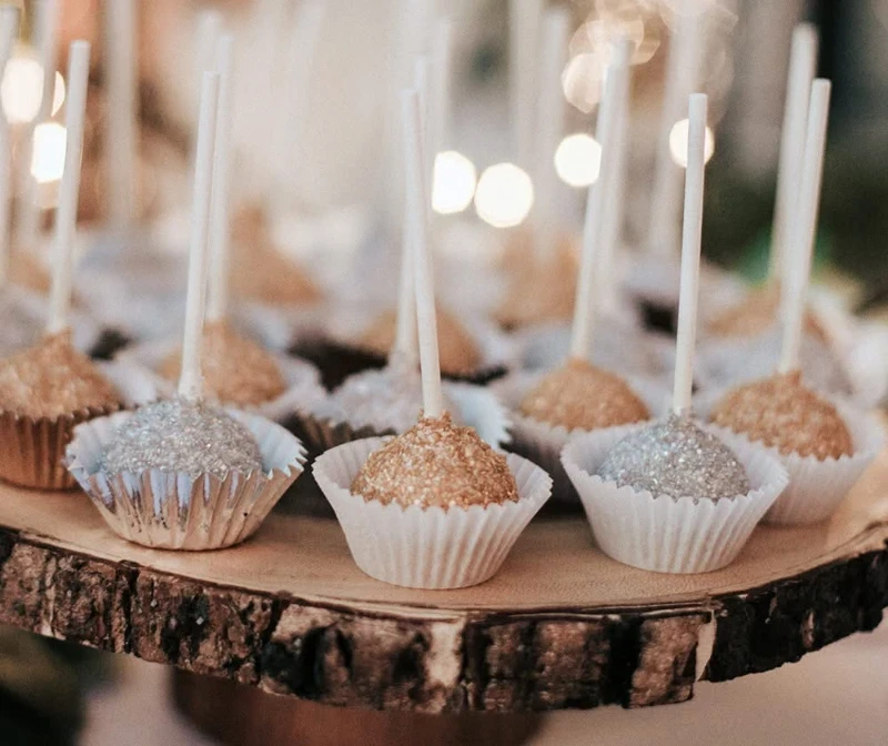 Cake pops coated with silver and gold glitter, displayed on a rustic wooden tray.
