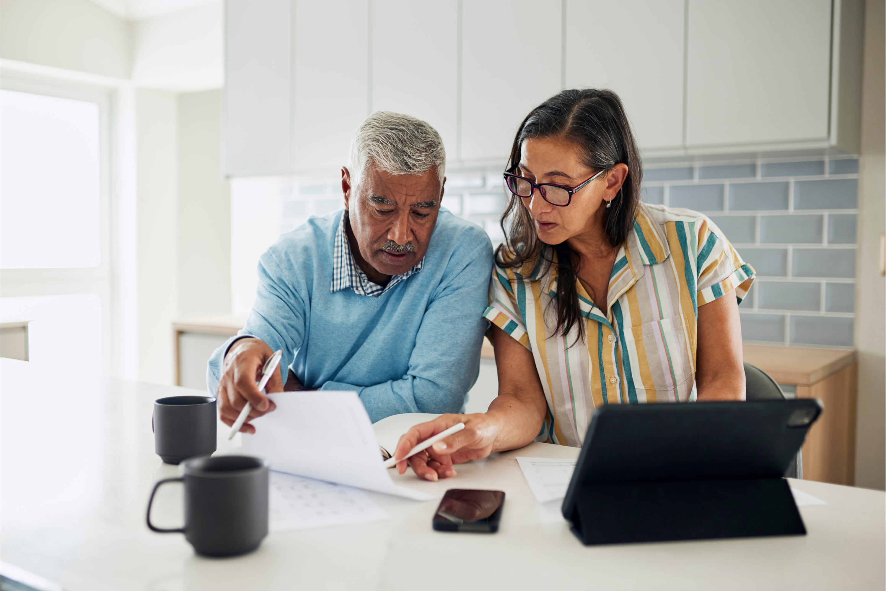 Older couple reviews documents together at a kitchen table with a tablet and coffee mugs.