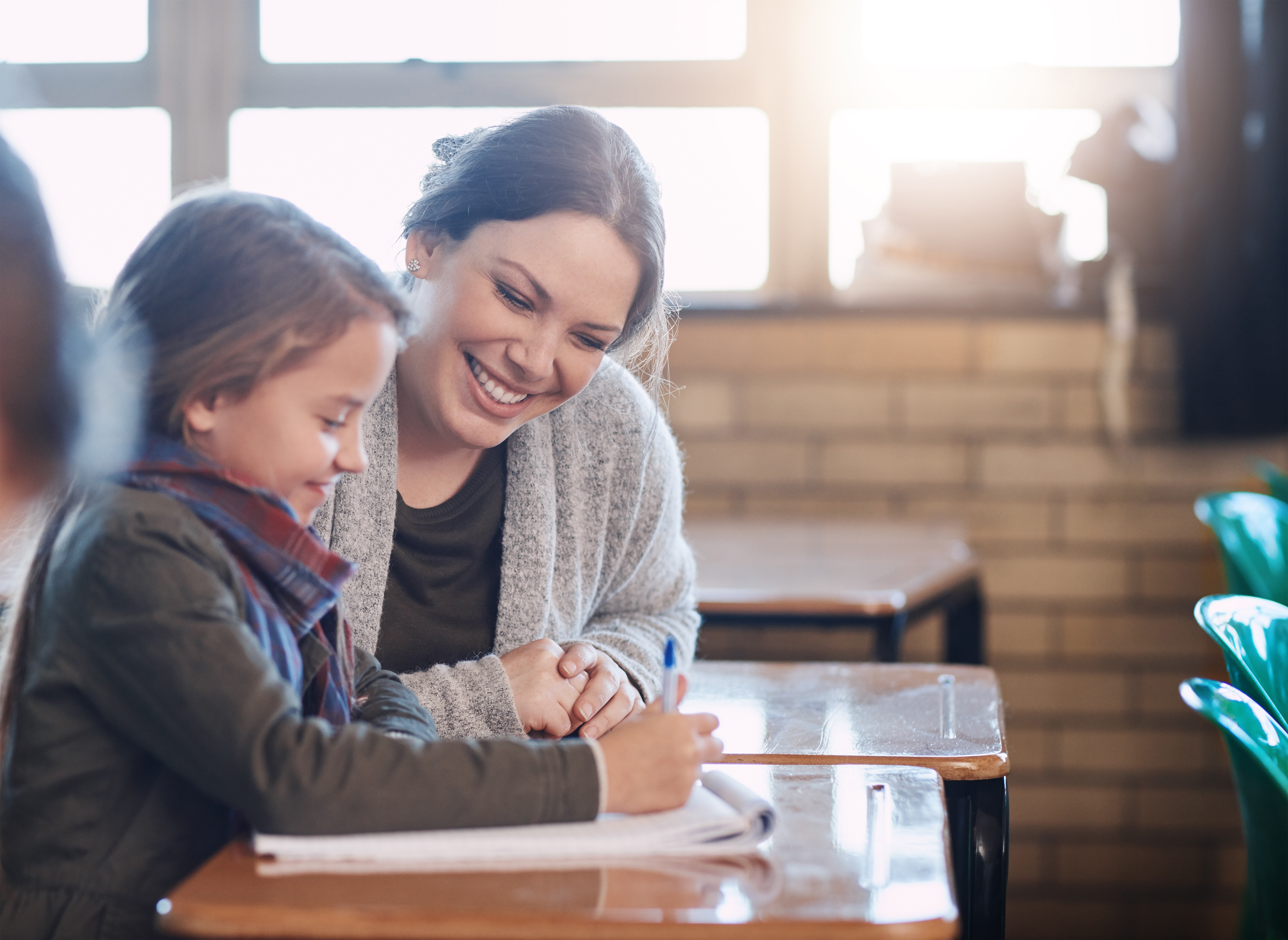 Smiling teacher helps a young student with schoolwork at a classroom desk by a window.