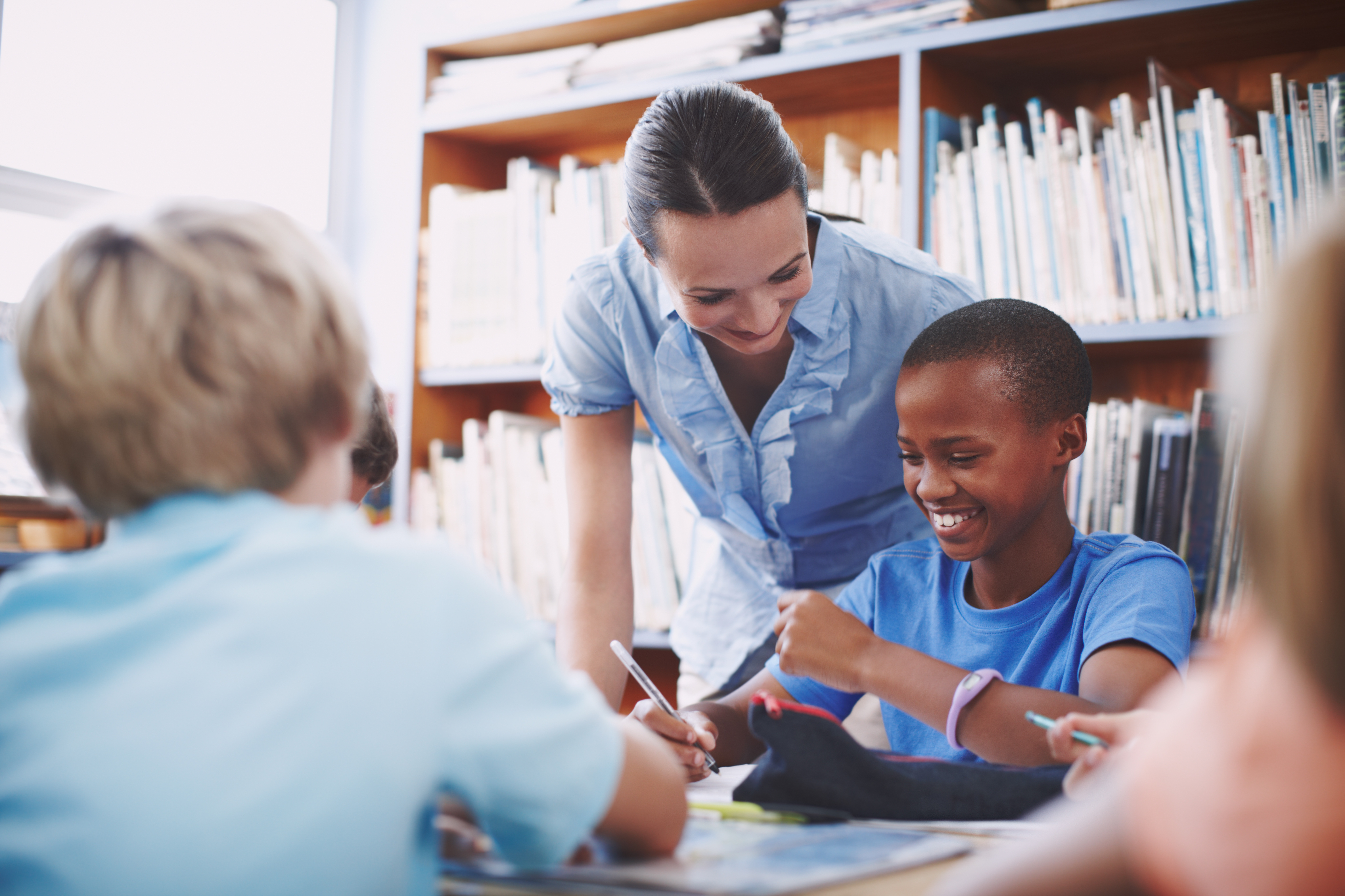 A teacher smiles while helping a student in a library with other kids at the table.