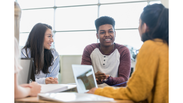 Three students sit at a table, talking and smiling, with laptops and notebooks in front of them.