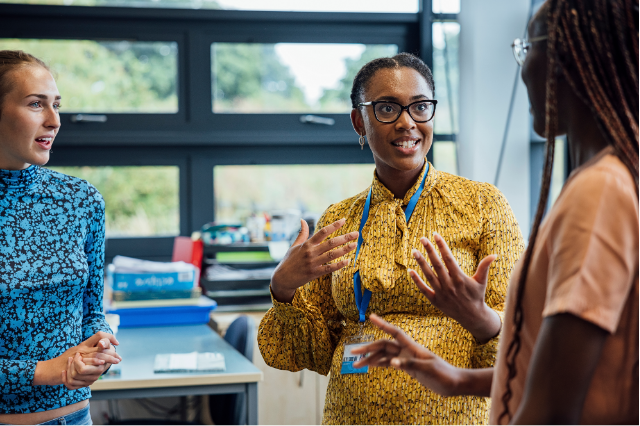 Three women standing and talking in an office, one gesturing with her hands while smiling.