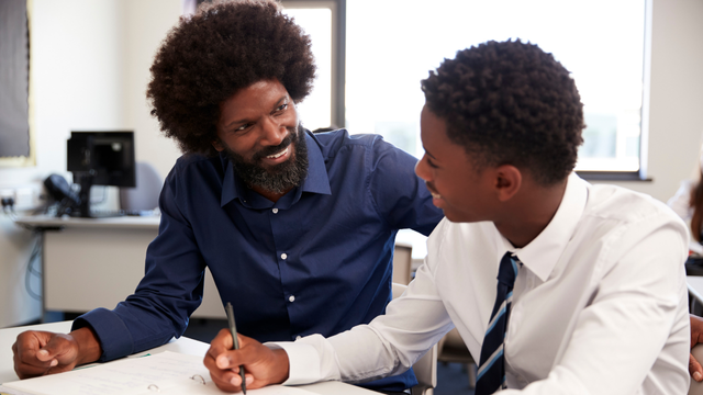 A teacher smiles and talks with a student in a classroom as they work together at a desk.