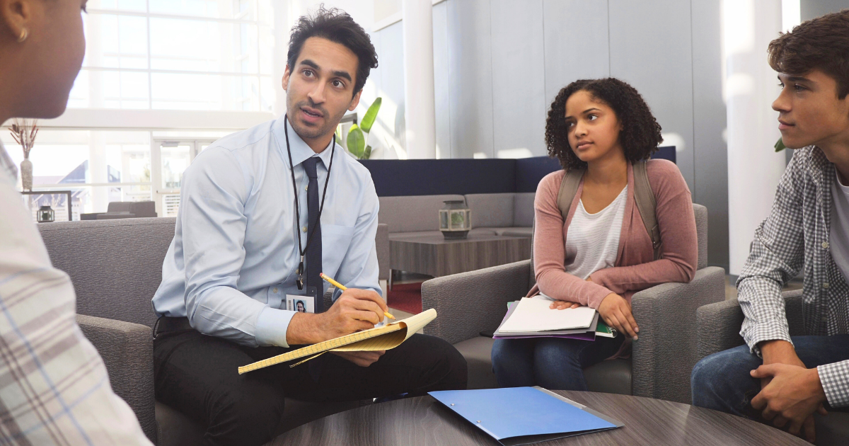 Man with notepad talks to two teens and another adult in a bright, modern office setting.