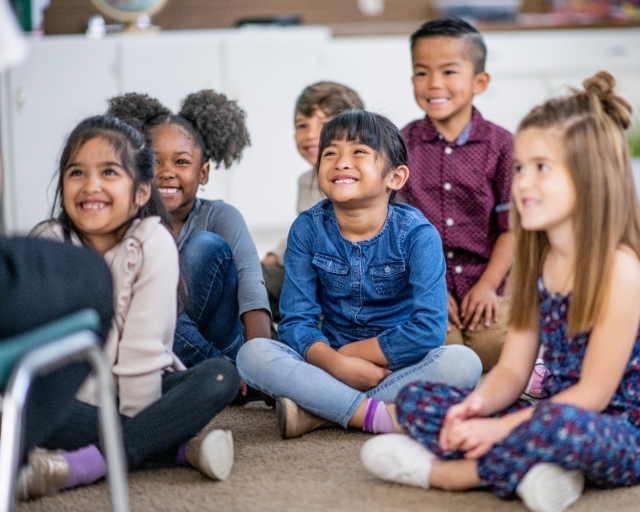 A group of young children sit cross-legged on the floor, smiling and listening attentively.