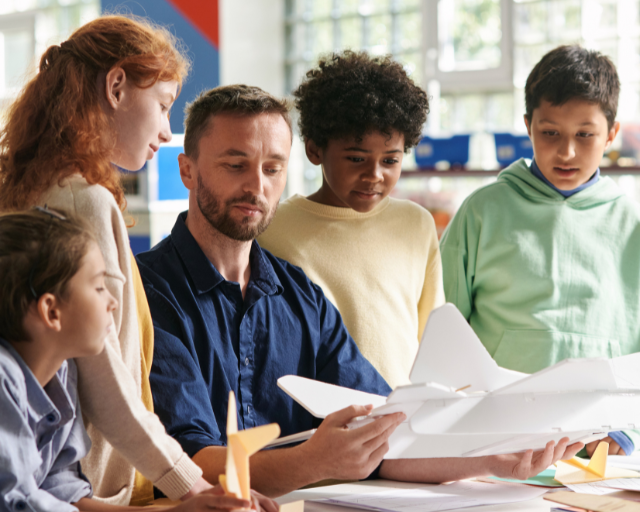 A teacher shows a model airplane to four attentive children in a bright classroom.