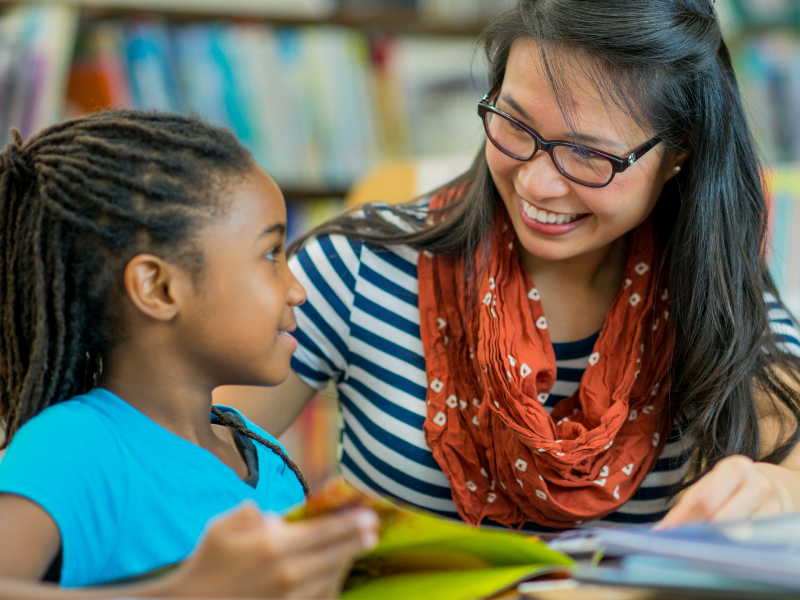 An adult and a child smile at each other while reading a book together in a library.