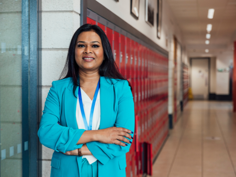A woman in a blue suit stands confidently with arms crossed in a school hallway by red lockers.
