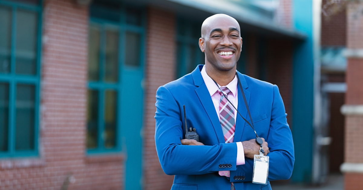 Smiling man in a blue suit with a lanyard and walkie-talkie standing outside a building.