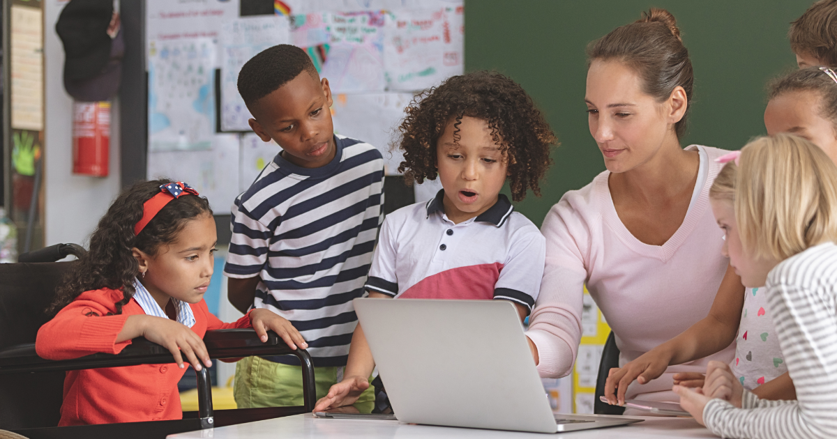 A long-term substitute teacher is sitting at a desk pointing to a computer screen. Six young students stand around the teacher, observing the screen.