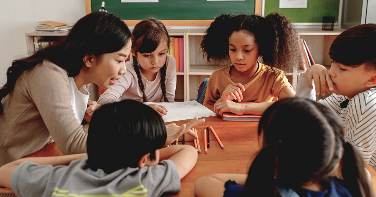 Smiling teacher holds clipboard; woman helps child at computer in a bright room.