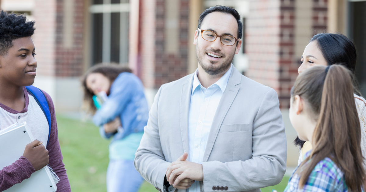 A smiling teacher stands outside talking with a group of students near a school building.