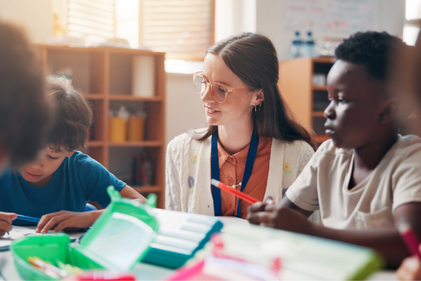 A teacher sits with two young students at a table, helping them with a classroom activity.
