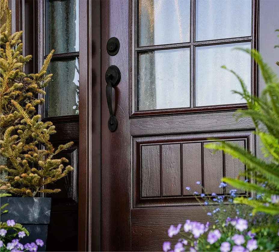 Close-up of a dark wooden front door with glass panels, surrounded by green plants and flowers.