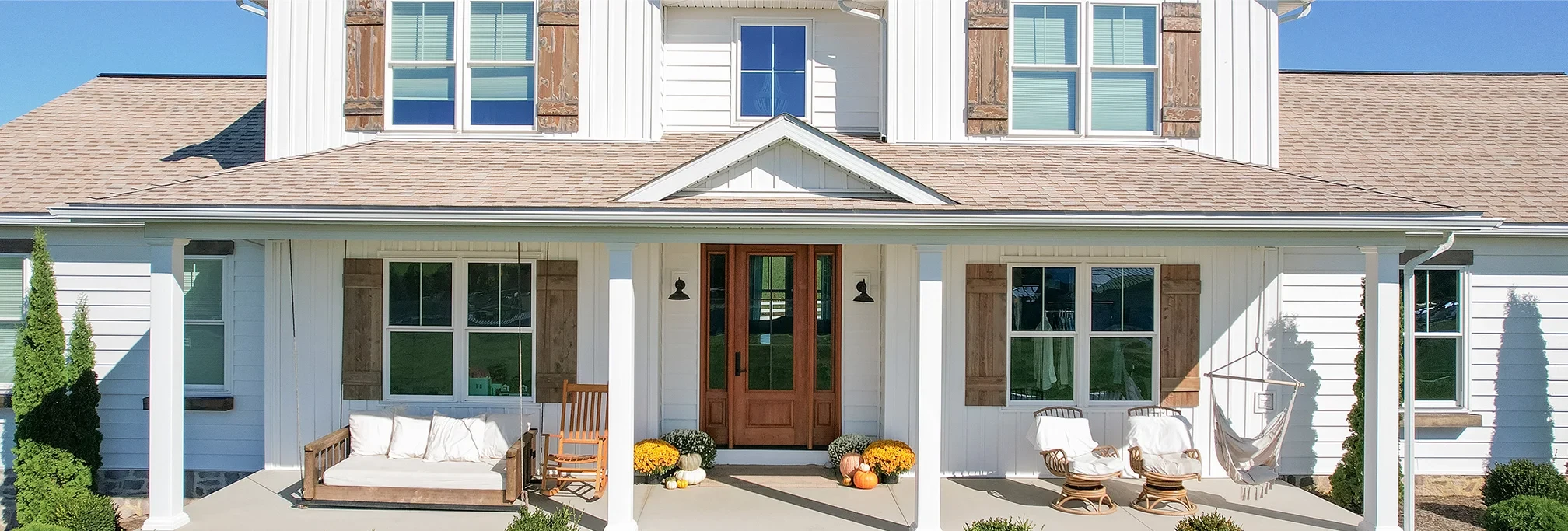 White house with wooden accents, front porch, chairs, bench, and potted yellow flowers.