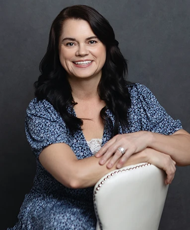 Woman with long dark hair in a blue dress smiling, sitting with arms on a white chair.