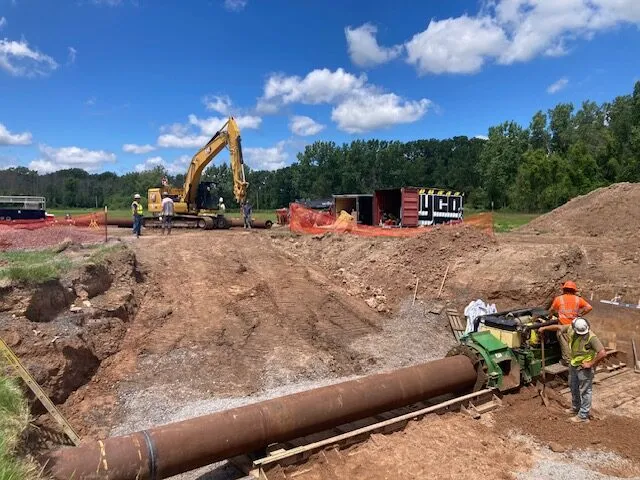 Construction workers operate heavy machinery and work on a large pipe at a dirt construction site.
