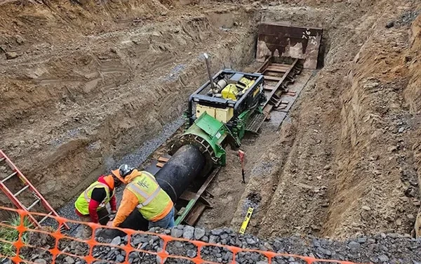Two workers install a large pipe underground at a construction site with heavy machinery nearby.