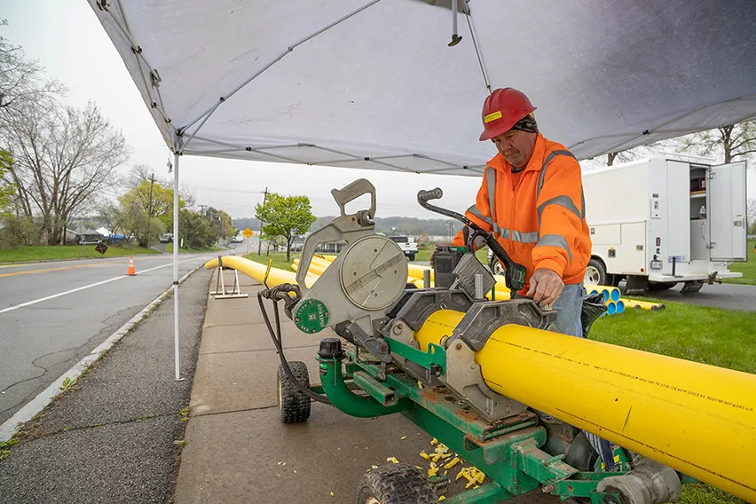 Workers in safety gear operate hoses on a snowy lawn near trucks and houses on a cold day.