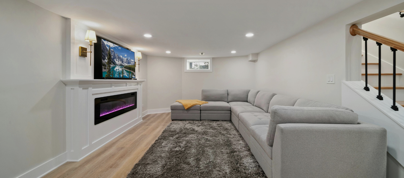 Modern basement living room with gray sectional sofa, rug, fireplace, and wall-mounted TV.