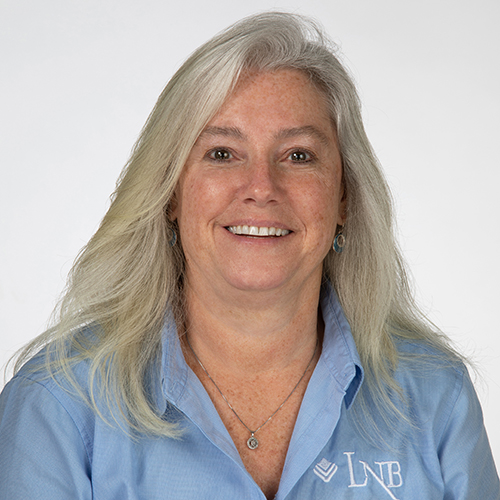 Smiling woman with long gray hair wearing a light blue LNB shirt against a white background.