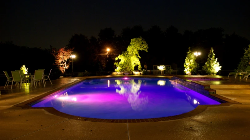 A swimming pool glows with purple lights at night, surrounded by trees and patio furniture.