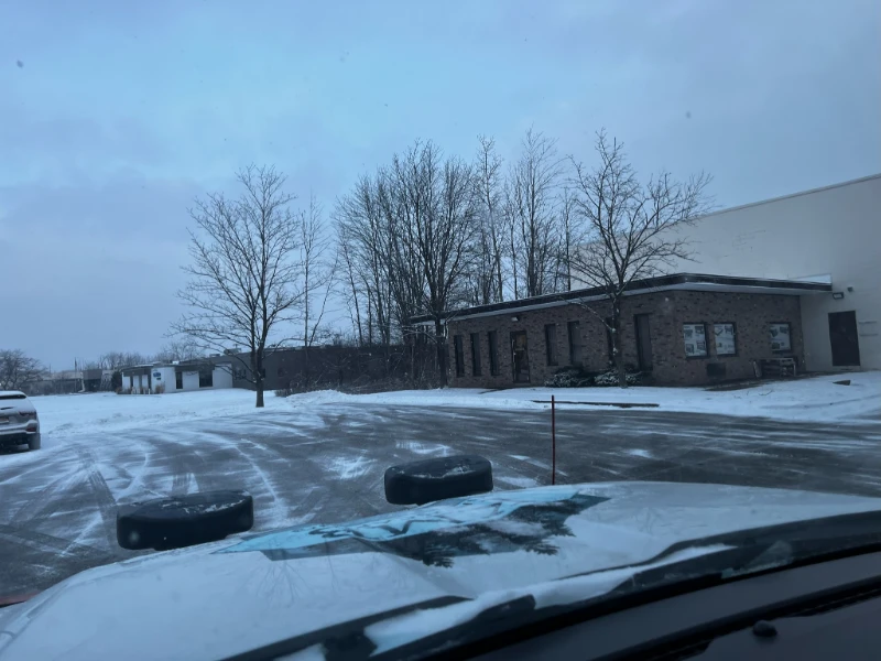 A snowy, empty parking lot in front of a brick building with bare trees in winter.