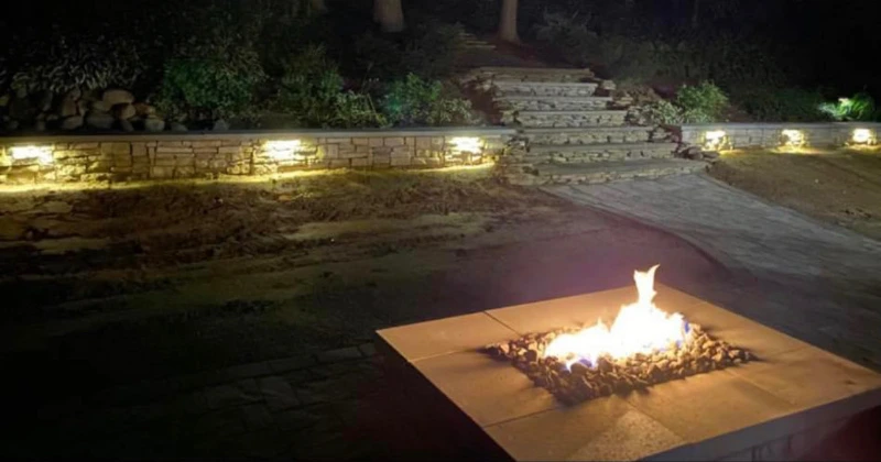 A square fire pit glows on a patio at night, with steps and garden lights in the background.