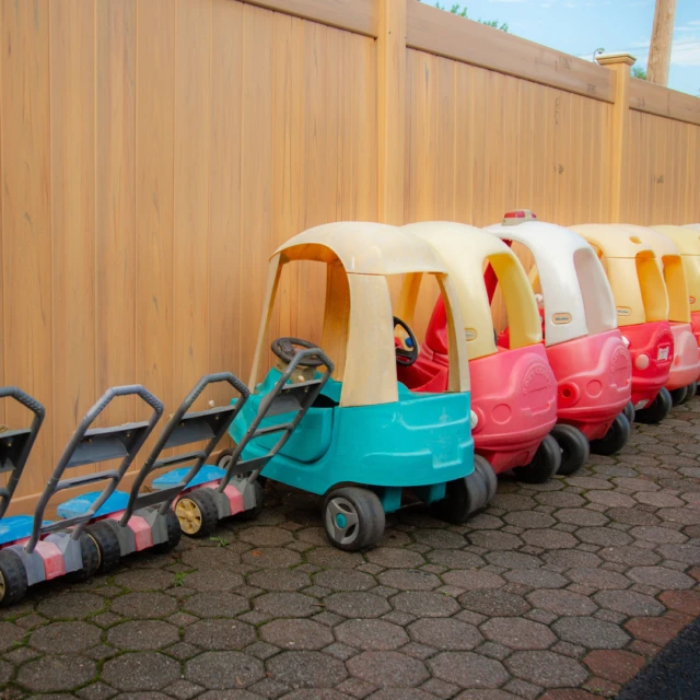 Toy cars and toy lawn mowers lined up on a sidewalk next to a tall wooden fence.