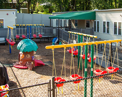 Outdoor playground with red swings, a playhouse, and wood chips, surrounded by fencing and buildings.