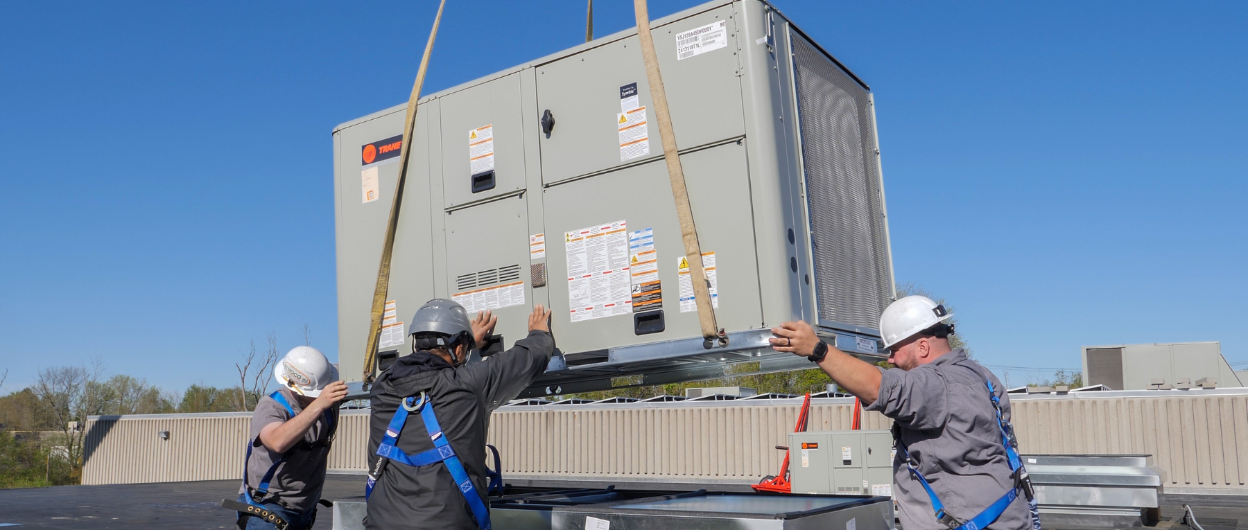 Two large outdoor HVAC units sit on a rooftop near pipes and a blue wooden wall.