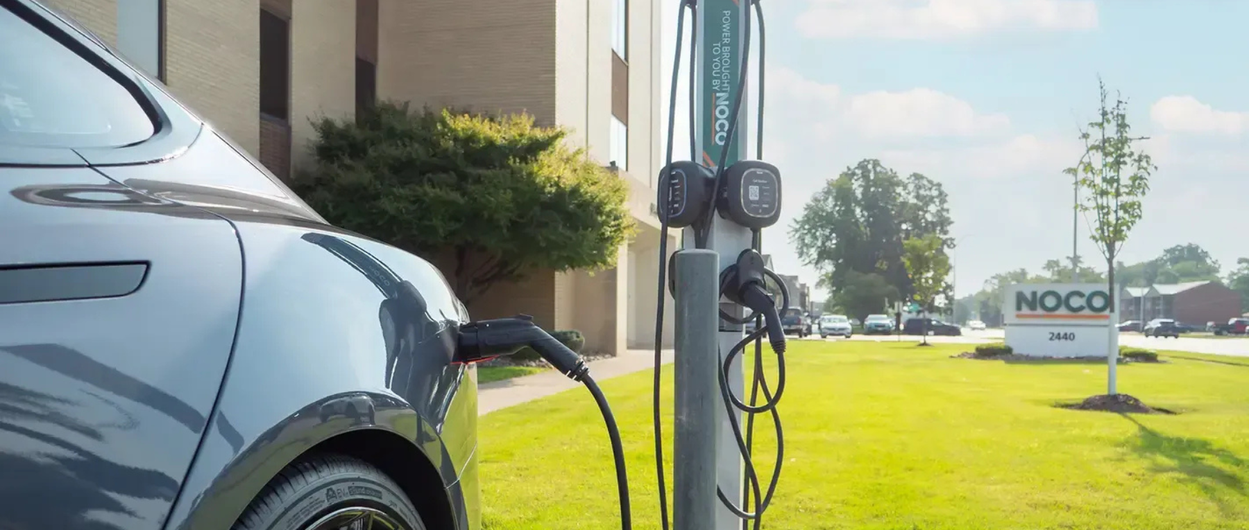 An electric car charging at a NOCO charging station outside a building on a sunny day.