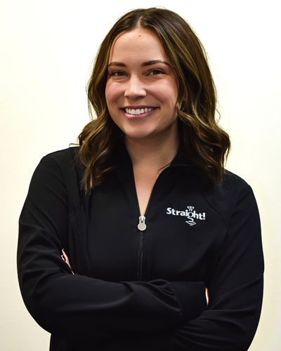 Woman with wavy brown hair smiling, wearing a black jacket with Straight logo, arms crossed.