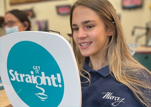 Young woman smiling at a dental mirror that says Get it Straight in a dental office.
