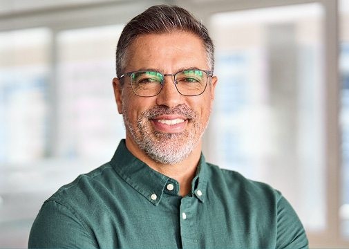 Smiling man with glasses and a green shirt standing indoors with blurred background.