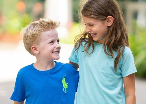 Smiling boy and girl in blue shirts standing close together outdoors, looking at each other.