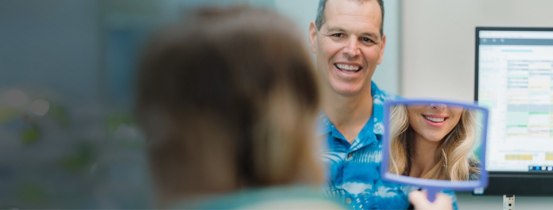 A woman smiles into a hand mirror, reflecting her mouth; a man smiles in the background.