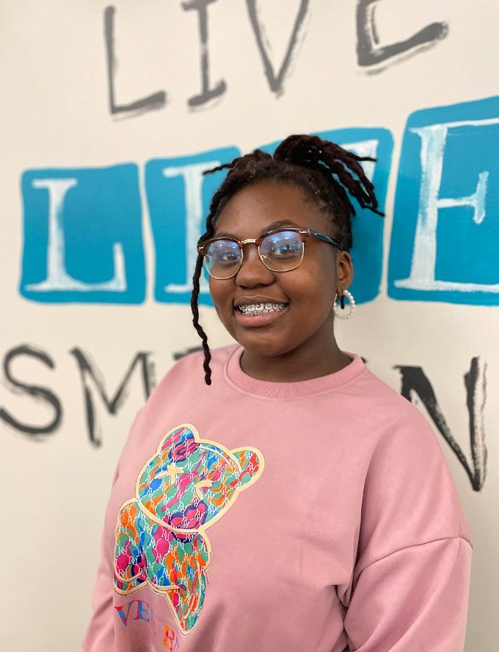 Smiling girl in glasses and pink bear sweatshirt stands before a wall reading LIVE LIFE SMILING.