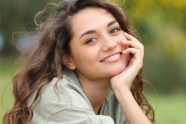 Woman with wavy brown hair smiling, resting her face on her hand outdoors in natural light.