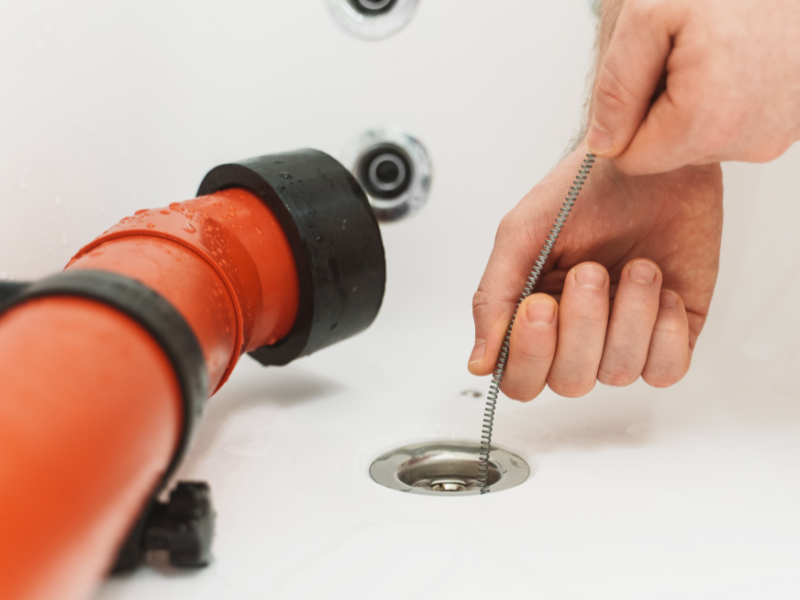 A person uses a metal drain snake to unclog a white sink with a red pipe visible.