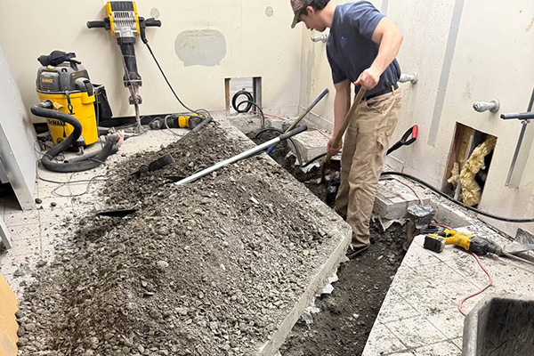 A worker in a cap digs up a tiled floor in a room under construction with tools nearby.
