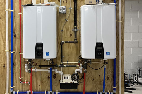 Two tankless water heaters mounted on a wooden wall with visible pipes and wiring.
